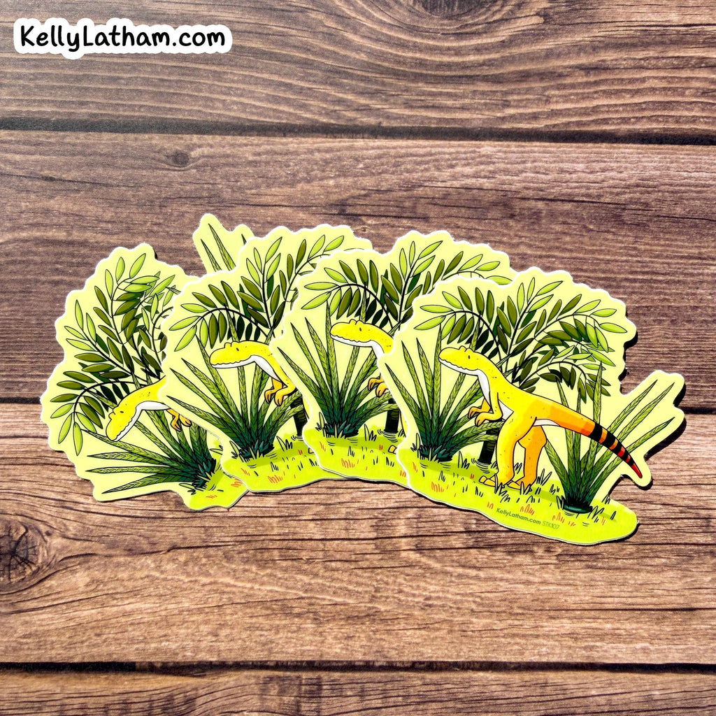 A die-cut vinyl sticker featuring a whimsical illustration of yellow lizards among green palm-like plants, placed on a brown wooden surface. The sticker has a white border and the brand name 'KellyLatham.