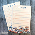 Two cream-colored to-do list notepads with blue lines and a colorful book-themed illustration at the bottom, displayed on a gray wood background.