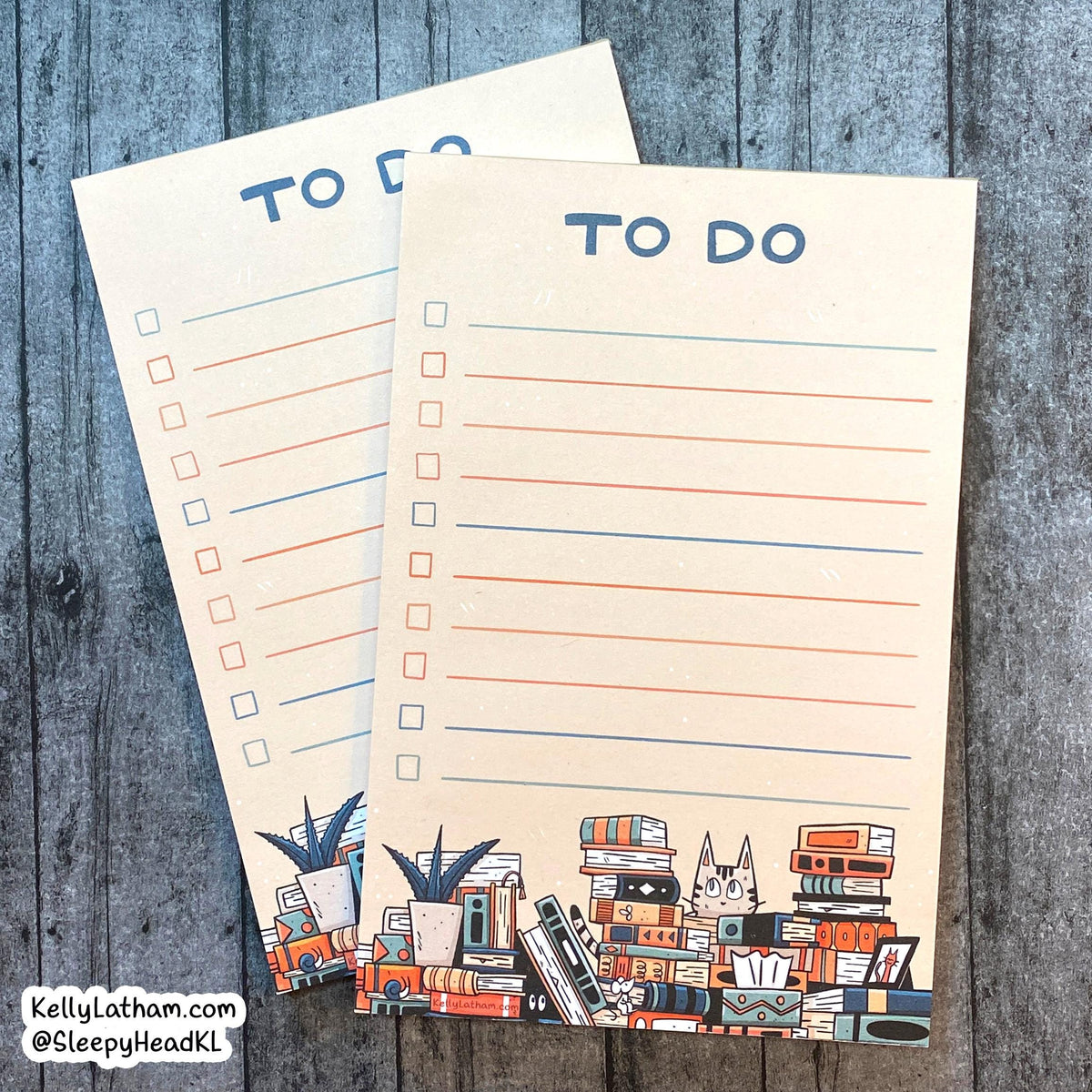 Two cream-colored to-do list notepads with blue lines and a colorful book-themed illustration at the bottom, displayed on a gray wood background.