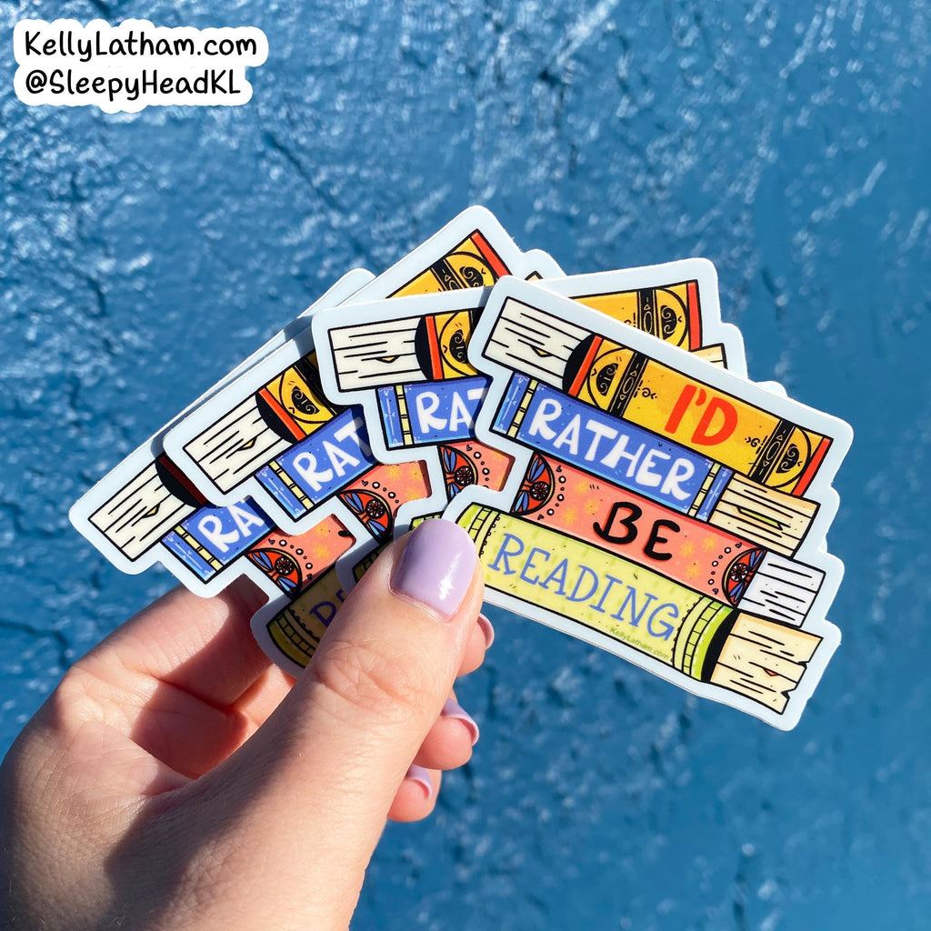 A hand with lavender nail polish holds a fan of colorful book-themed stickers against a bright blue textured background. The stickers feature illustrations of stacked books with phrases like 'RATHER BE READING' and 'I'D BE READING'.