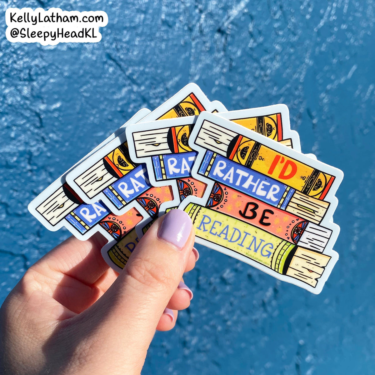A hand with lavender nail polish holds a fan of colorful book-themed stickers against a bright blue textured background. The stickers feature illustrations of stacked books with phrases like 'RATHER BE READING' and 'I'D BE READING'.