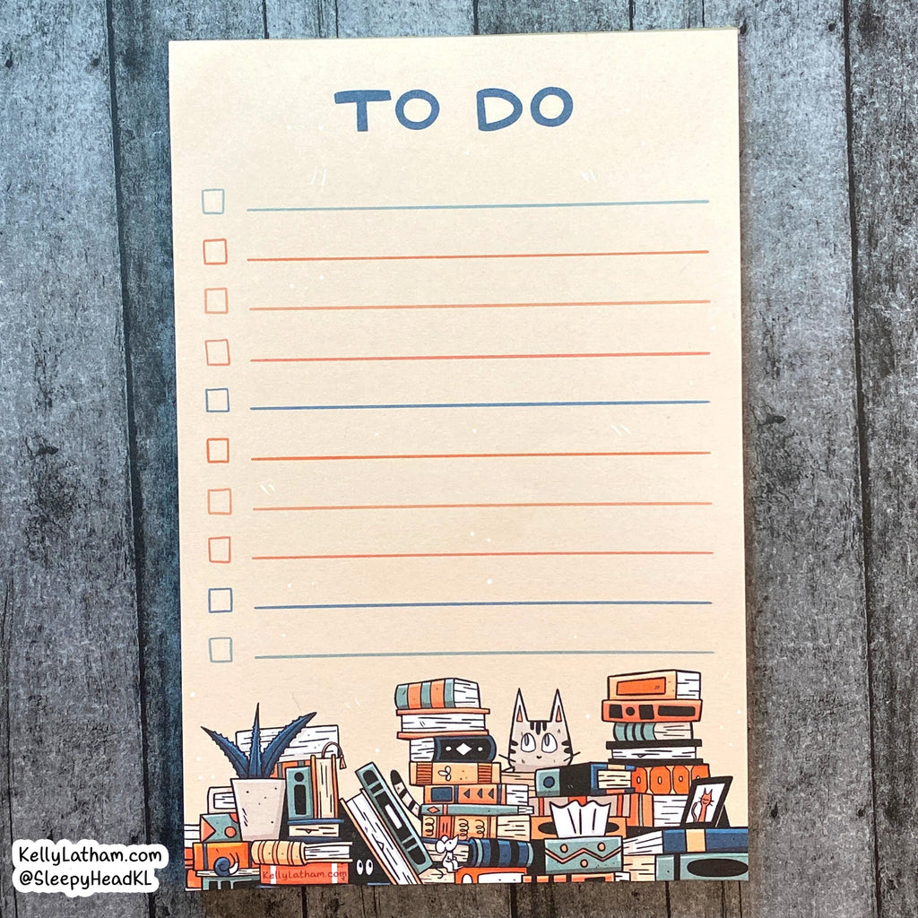 A vertical cream-colored notepad with a 'TO DO' checklist layout, featuring blue headers, orange and blue lines, and an illustration of stacked books, a plant, and a cat at the bottom. The notepad is photographed flat on a gray wood plank background.