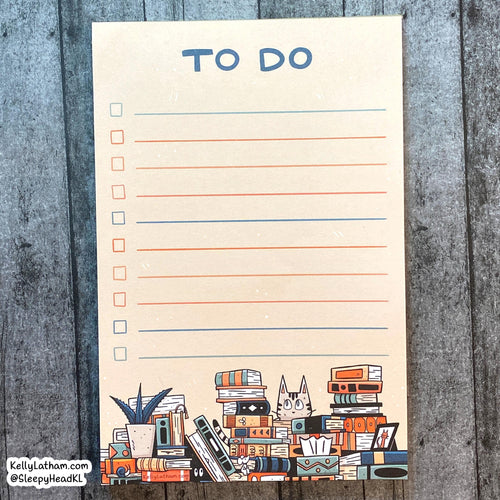 A vertical cream-colored notepad with a 'TO DO' checklist layout, featuring blue headers, orange and blue lines, and an illustration of stacked books, a plant, and a cat at the bottom. The notepad is photographed flat on a gray wood plank background.
