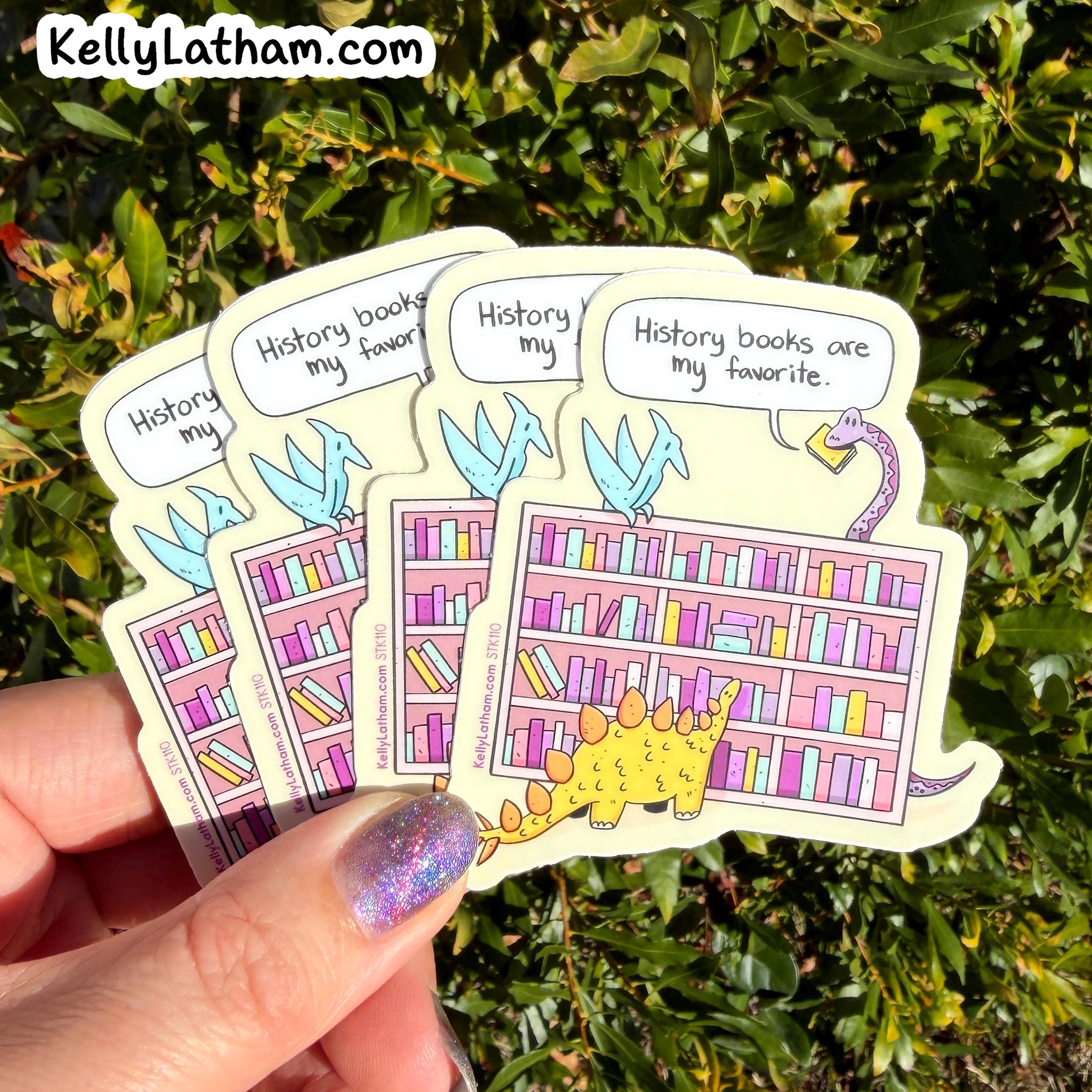 A hand with glittery purple nails holds a fan of four die-cut stickers featuring cartoon dinosaurs and bookshelves with the phrase 'History books are my favorite.' The background is green foliage.