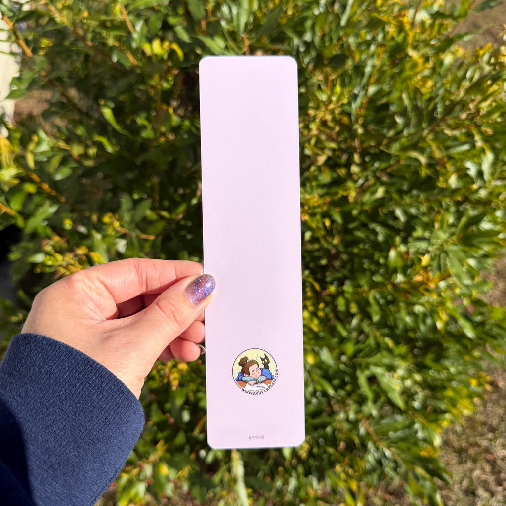 A hand holding a long, narrow light lavender card or bookmark with a small circular dog illustration near the bottom, set against a blurred background of green foliage.