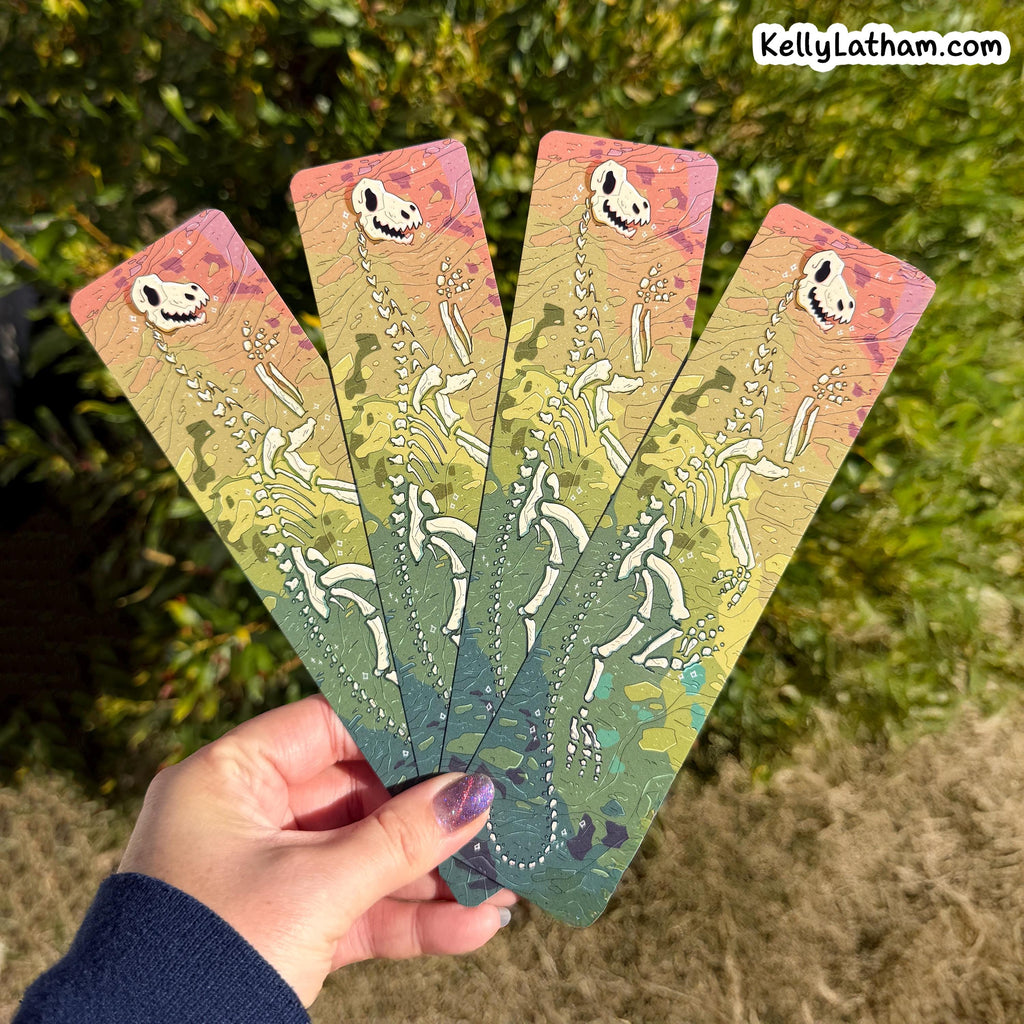 A hand holds four long rectangular bookmarks or art cards fanned out against an outdoor background of green foliage. Each card features a white skeleton figure on a pastel gradient background transitioning from pink and peach at the top to yellow and green at the bottom.