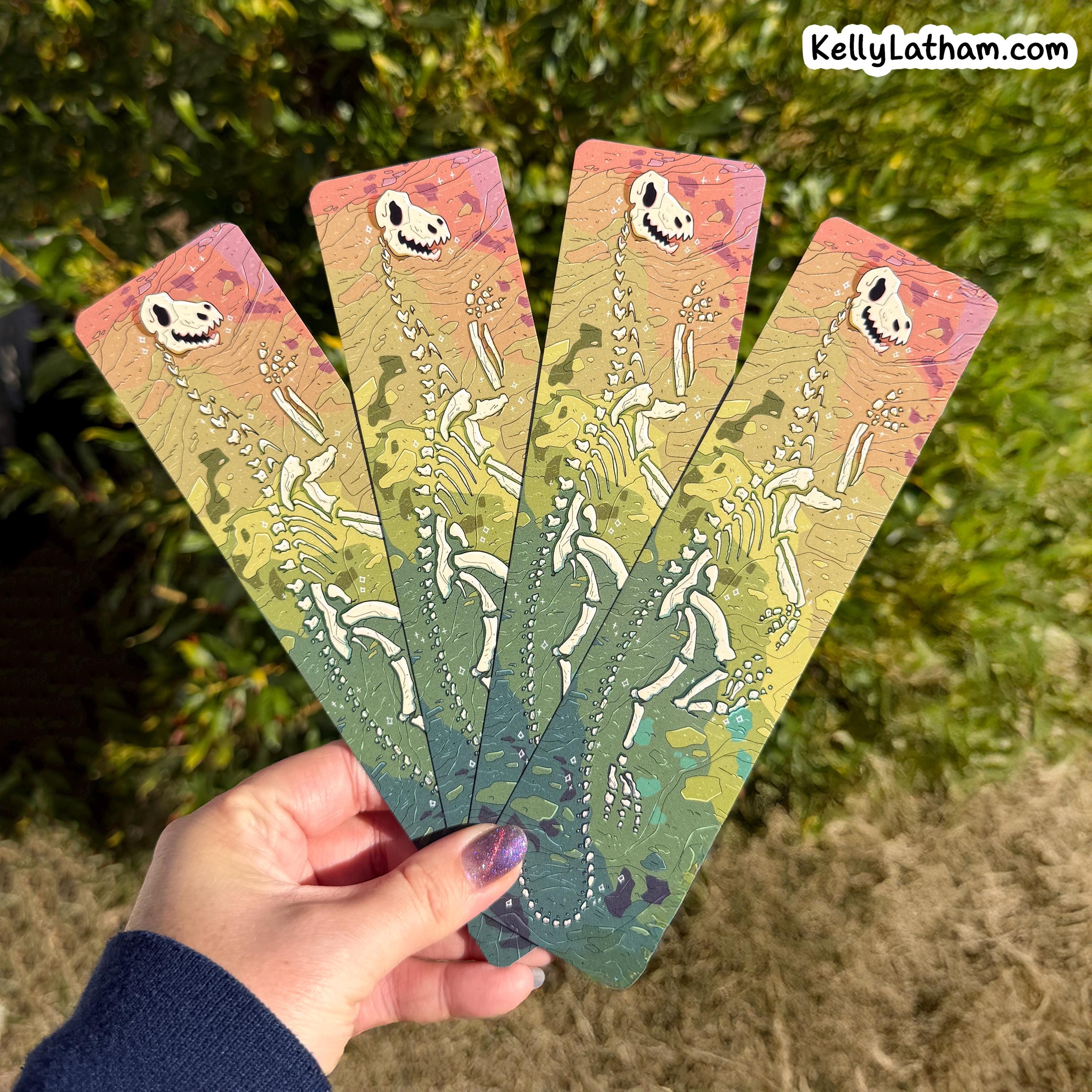 A hand holds four long rectangular bookmarks or art cards fanned out against an outdoor background of green foliage. Each card features a white skeleton figure on a pastel gradient background transitioning from pink and peach at the top to yellow and green at the bottom.