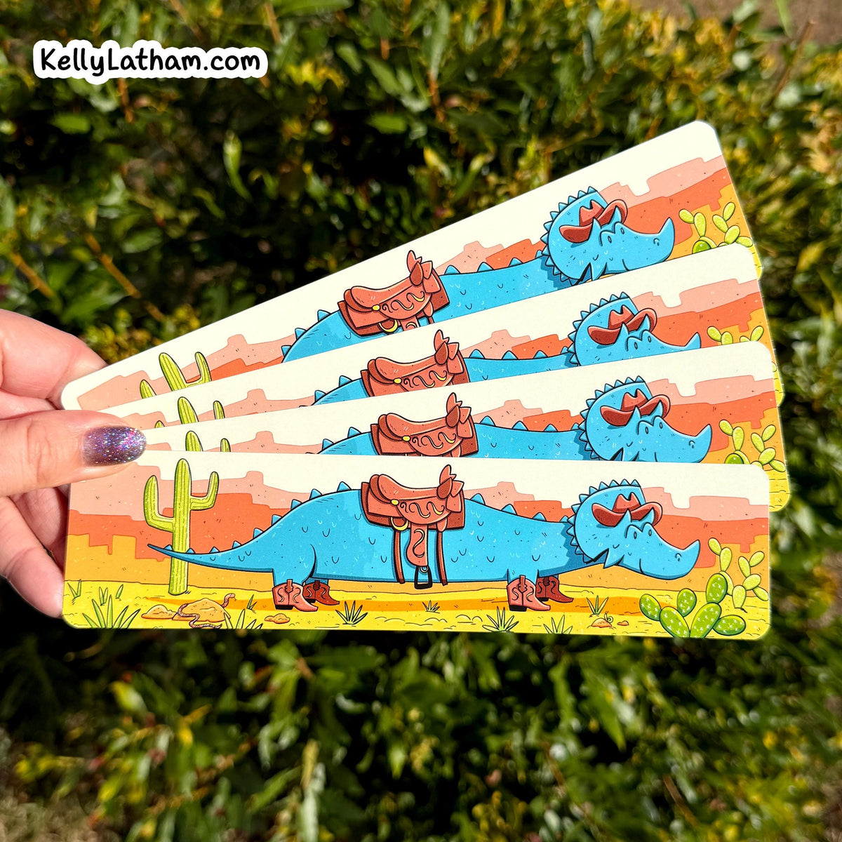 A hand holds four rectangular printed bookmarks featuring a cartoon blue dinosaur wearing a brown cowboy saddle and boots in a desert landscape, with green foliage in the background. The top-left corner shows the text 'KellyLatham.
