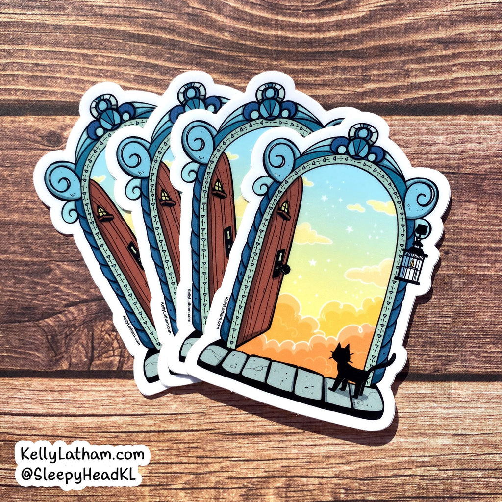 Three illustrated die-cut stickers showing ornate blue doorways opening to a sunset sky, arranged on a wood surface. A small black cat silhouette stands at the bottom of one doorway.