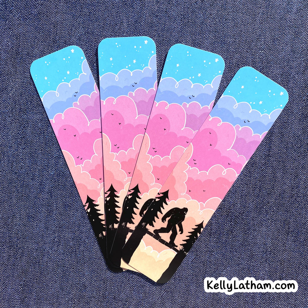 Four matching bookmarks fanned out on a blue denim background, featuring a pastel sky with pink, purple, and blue clouds, stars, and tree silhouette of bigfoot plus a Bigfoot silhouette on the bottom.