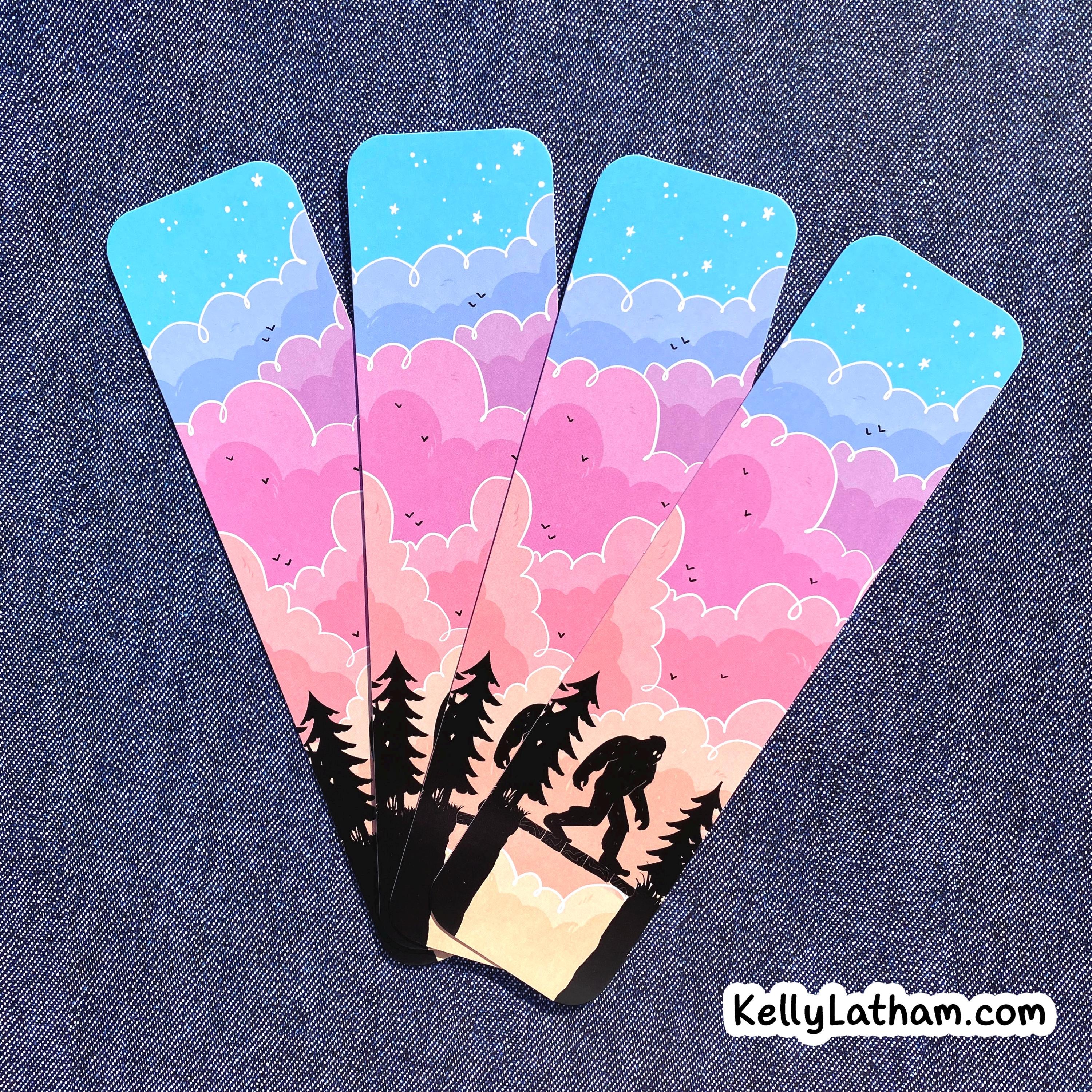 Four matching bookmarks fanned out on a blue denim background, featuring a pastel sky with pink, purple, and blue clouds, stars, and tree silhouette of bigfoot plus a Bigfoot silhouette on the bottom.