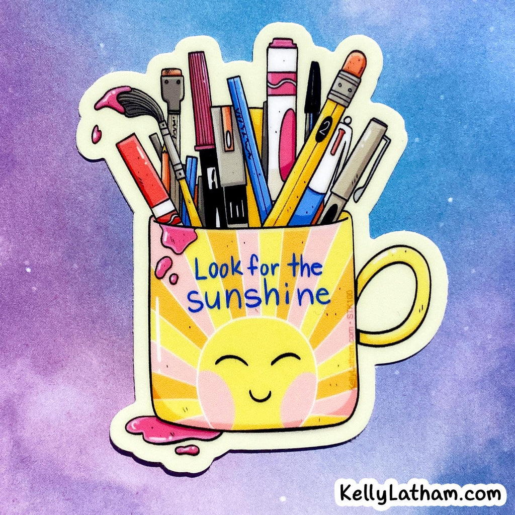 A die-cut sticker featuring a yellow sun face inside a mug filled with colorful art supplies, with the text 'Look for the sunshine' on the mug, shown on a purple and blue watercolor background.