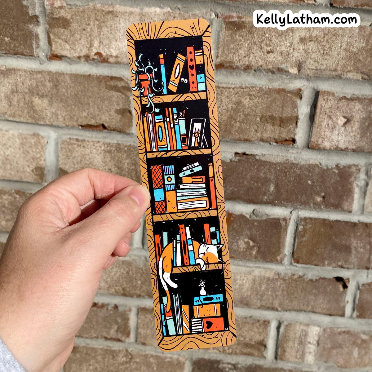 A hand holds a long rectangular illustrated bookmark or card featuring stylized bookshelves, books, and a cat in orange, black, blue, and cream tones, against a brick wall background. The text 'KellyLatham.