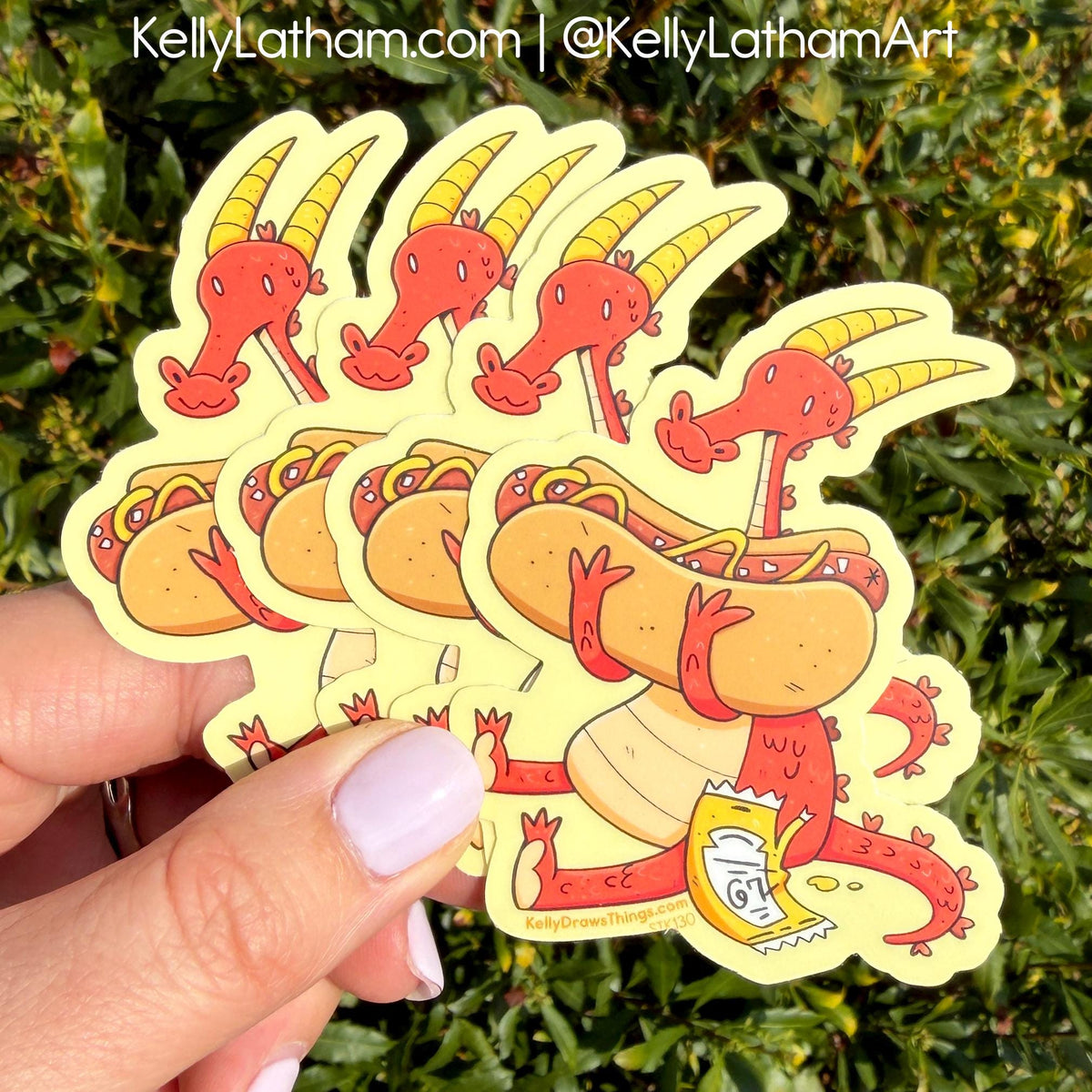 A hand holds a fan of die-cut stickers featuring a red cartoon dragon carrying a hot dog, with green foliage in the background. The text 'KellyLatham.
