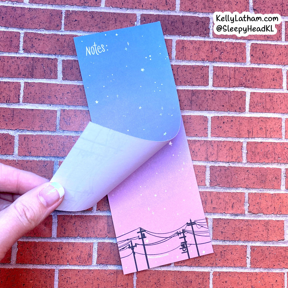 A hand peels back the top sheet of a pastel gradient notepad with a starry sky design, set against a red brick wall background. The bottom sheet shows a black line drawing of power lines.
