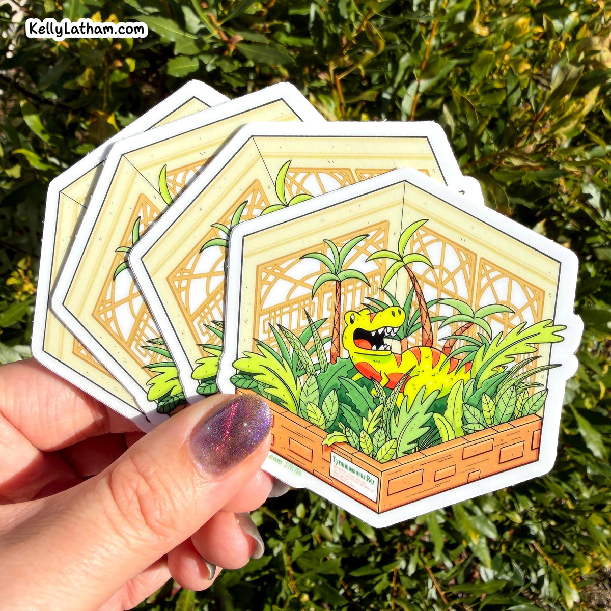 A hand holds a fan of hexagonal stickers featuring a cartoon green dinosaur in a jungle scene, with a real outdoor leafy background behind it.