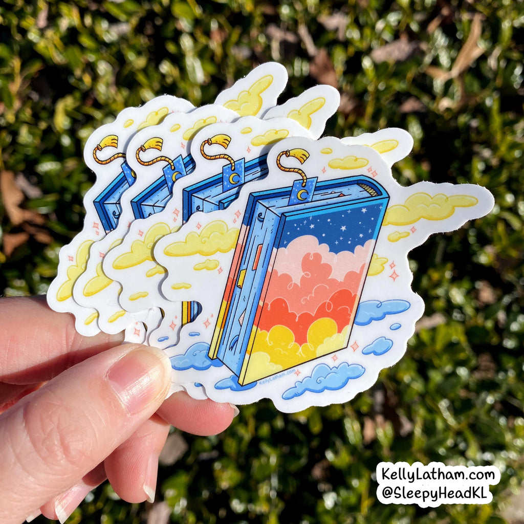 A hand holds a stack of die-cut stickers featuring an illustrated open book with a colorful sunset sky inside, set against a blurred green outdoor background. A watermark in the bottom right reads KellyLatham.