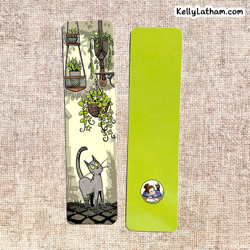 Two decorative paper bookmarks laid flat on a beige woven fabric background. The left bookmark is illustrated with hanging plants, a gray cat, and a cobblestone path.
