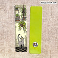 Two decorative paper bookmarks laid flat on a beige woven fabric background. The left bookmark is illustrated with hanging plants, a gray cat, and a cobblestone path.