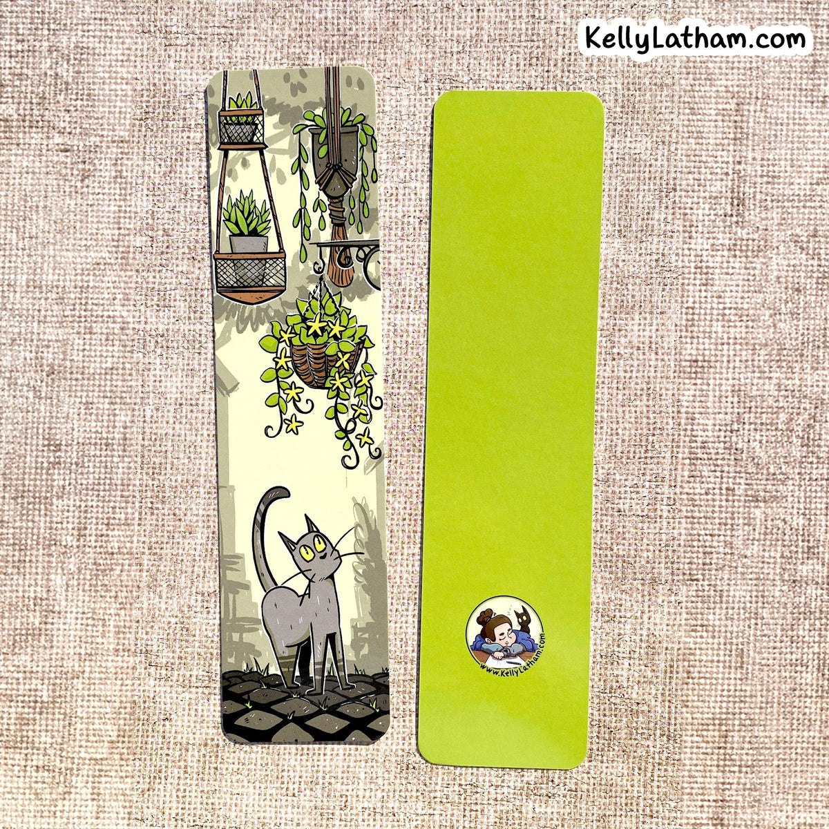 Two decorative paper bookmarks laid flat on a beige woven fabric background. The left bookmark is illustrated with hanging plants, a gray cat, and a cobblestone path.