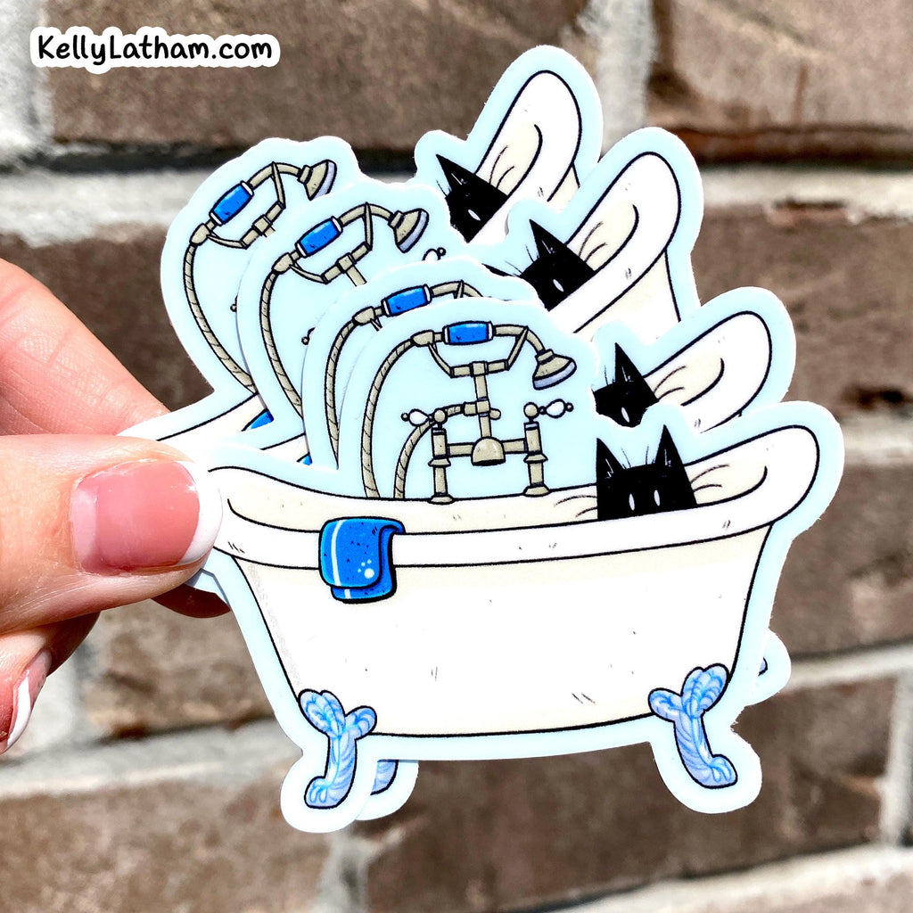 A hand holds a die-cut sticker featuring a white clawfoot bathtub filled with multiple bathtubs, showerheads, hoses, and black-and-white cat heads, against a blurred brown brick wall. The sticker has a light blue outline and a blue towel detail.
