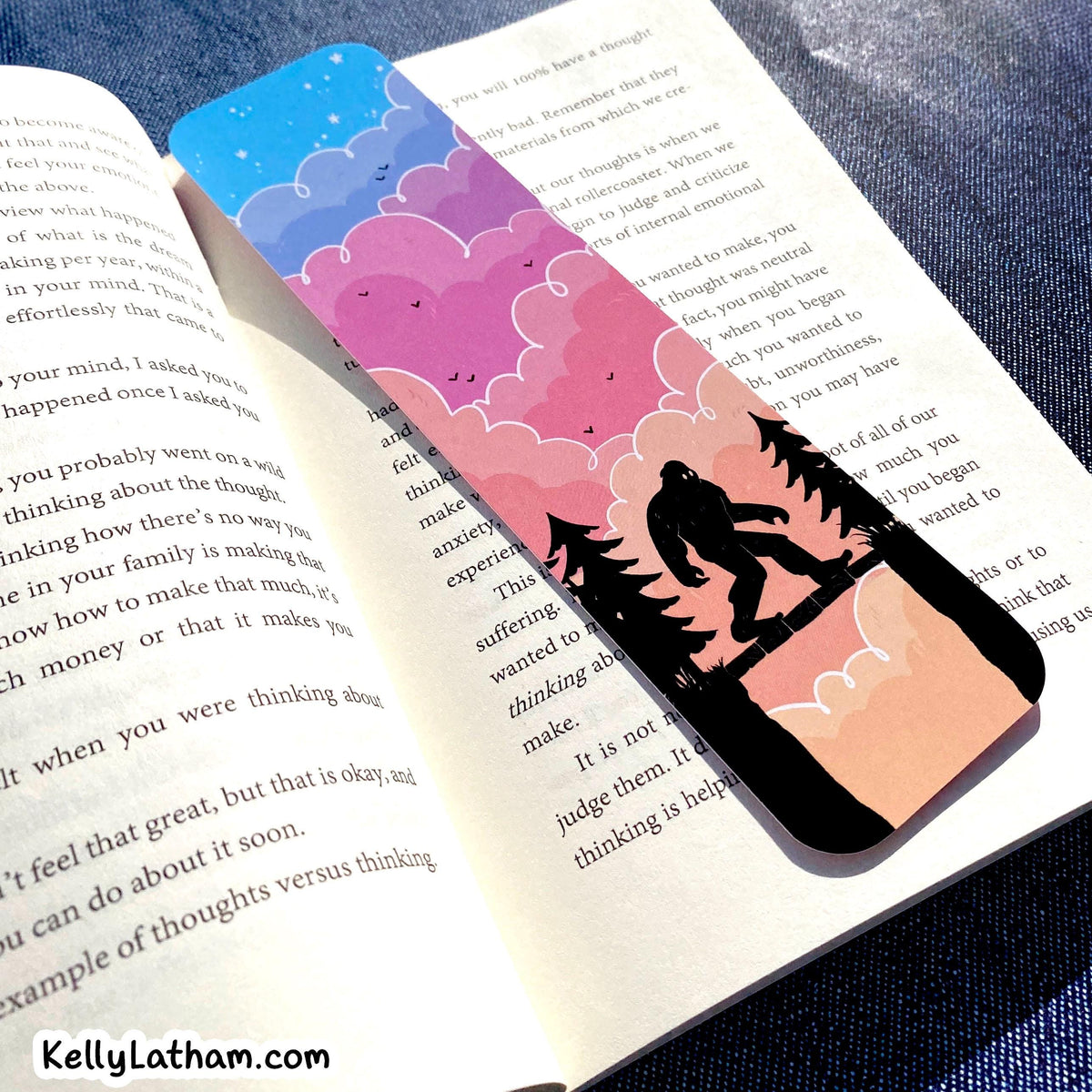 A rectangular bookmark with a colorful illustration of a Bigfoot silhouette standing in front of pink and purple clouds and a blue starry sky, placed on open book pages.