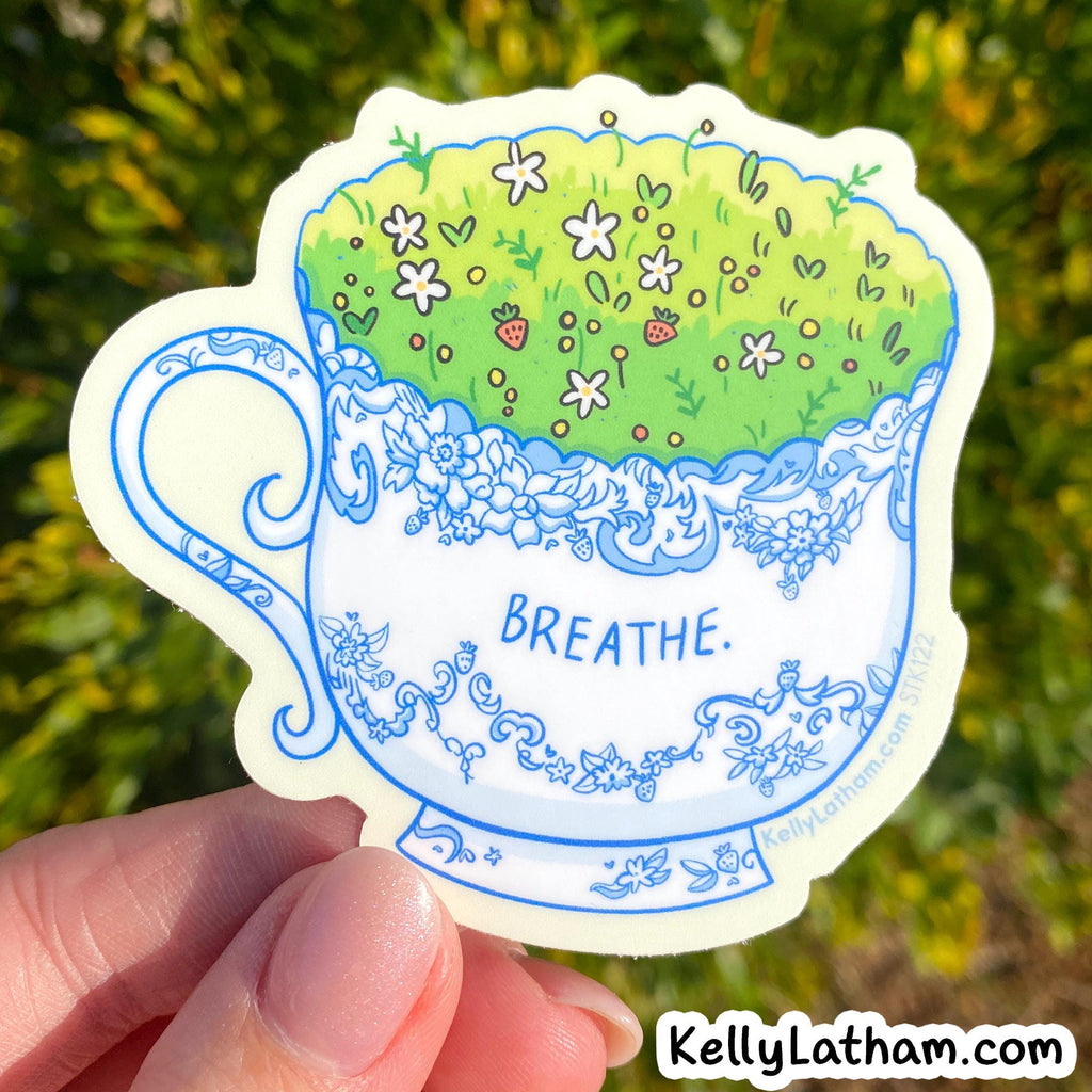 A hand holds a die-cut sticker shaped like a white teacup with blue floral detailing and a green grassy top covered in small white flowers and a strawberry. The word “BREATHE.