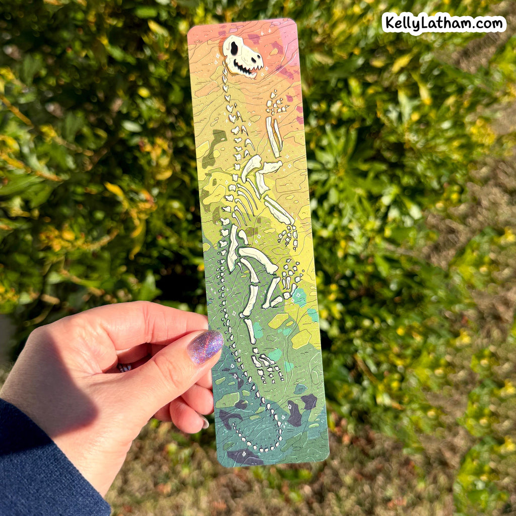 A hand holds a long rectangular printed card featuring a dinosaur skeleton illustration over a pastel gradient background with green foliage outdoors behind it. A small 'KellyLatham.