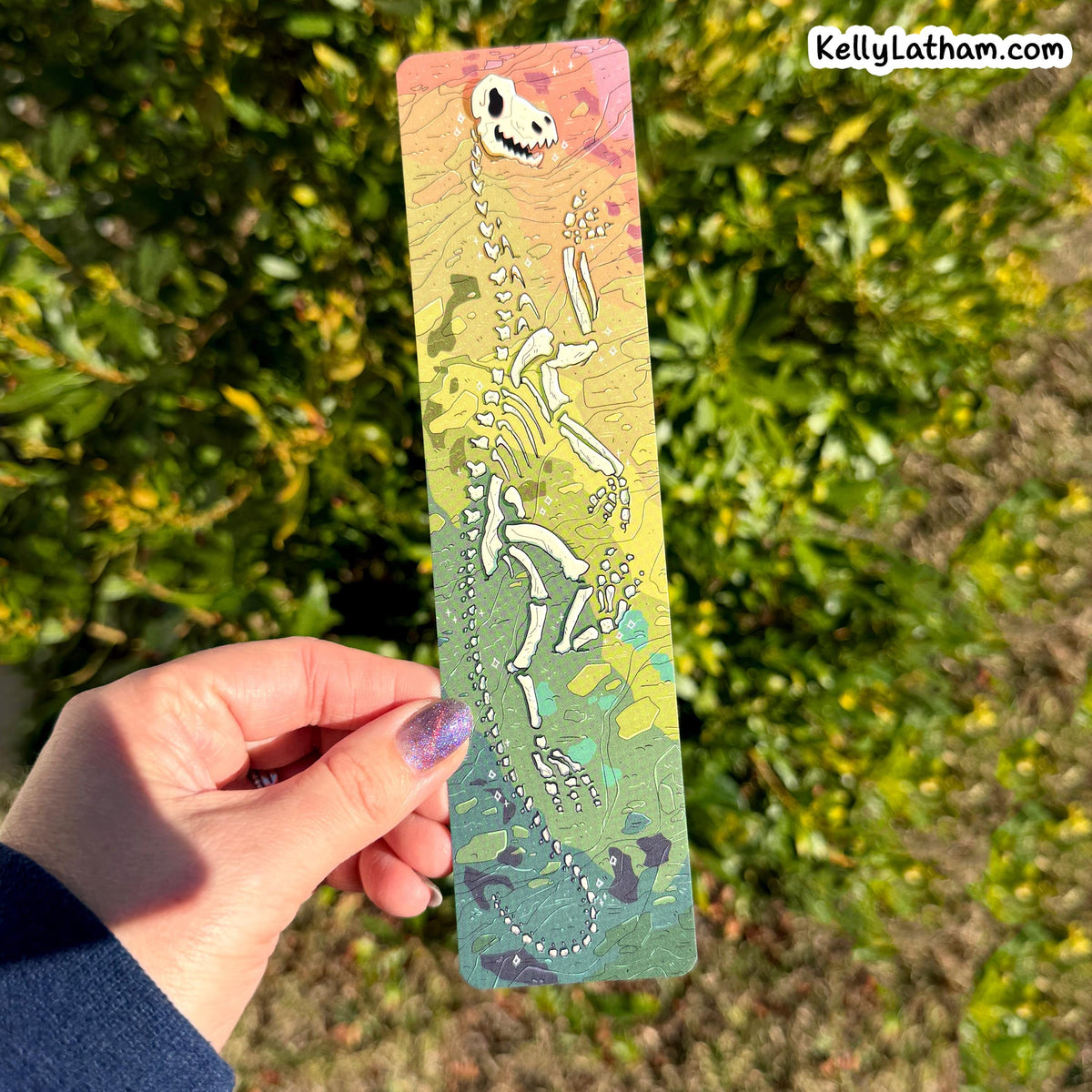 A hand holds a long rectangular printed card featuring a dinosaur skeleton illustration over a pastel gradient background with green foliage outdoors behind it. A small 'KellyLatham.