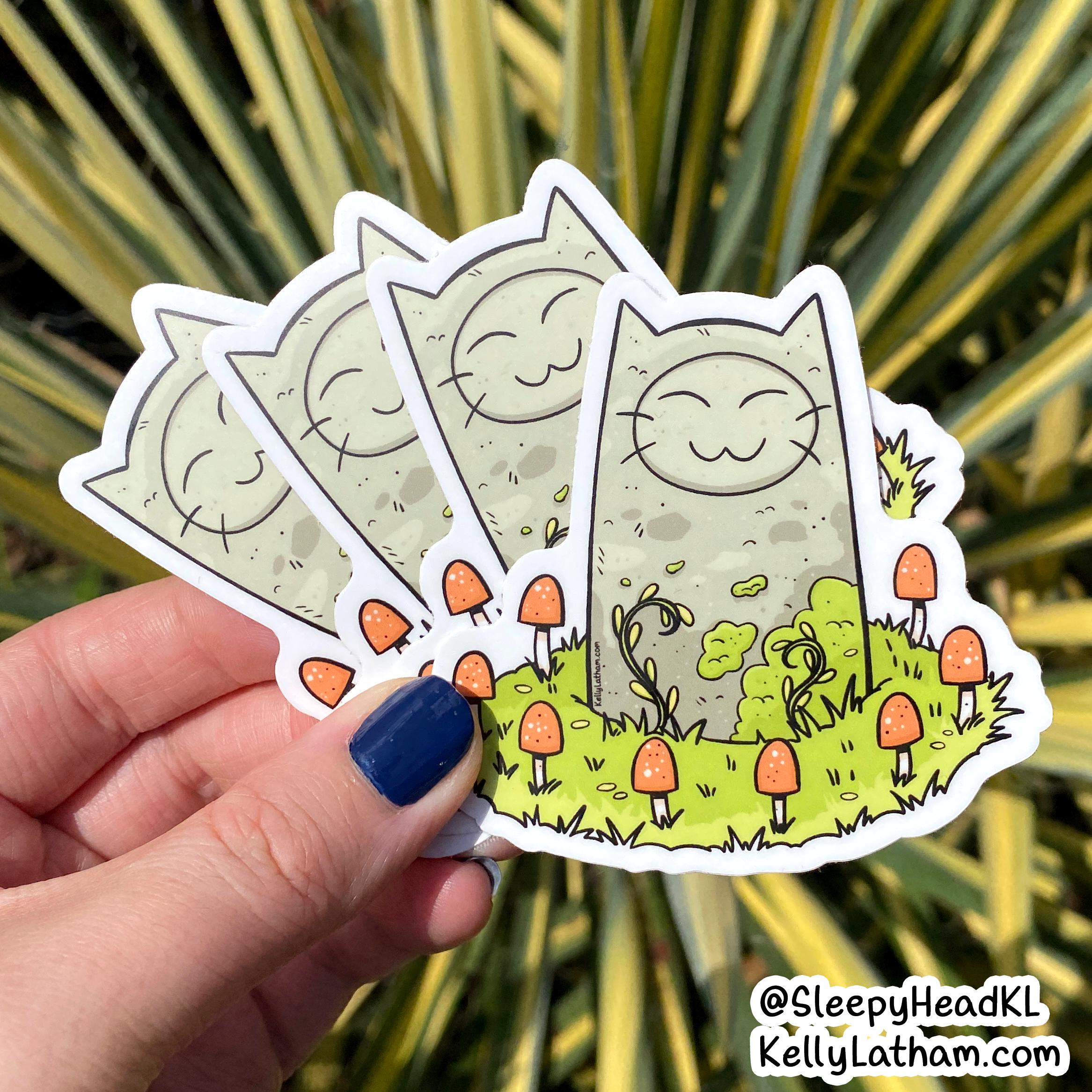 A hand with dark blue nail polish holds three die-cut stickers featuring a cute cartoon cat in a grassy field with orange mushrooms, set against a background of green and yellow variegated plant leaves.