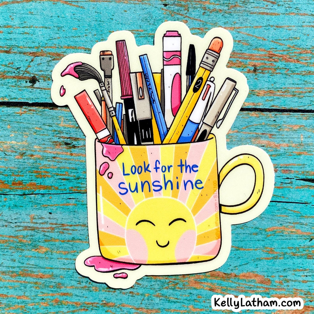 A die-cut sticker featuring a yellow and pink sun-faced mug filled with colorful art supplies, placed on a distressed turquoise wooden surface. The sticker reads 'Look for the sunshine' and includes a small 'Kellylatham.