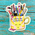 A die-cut sticker featuring a yellow and pink sun-faced mug filled with colorful art supplies, placed on a distressed turquoise wooden surface. The sticker reads 'Look for the sunshine' and includes a small 'Kellylatham.