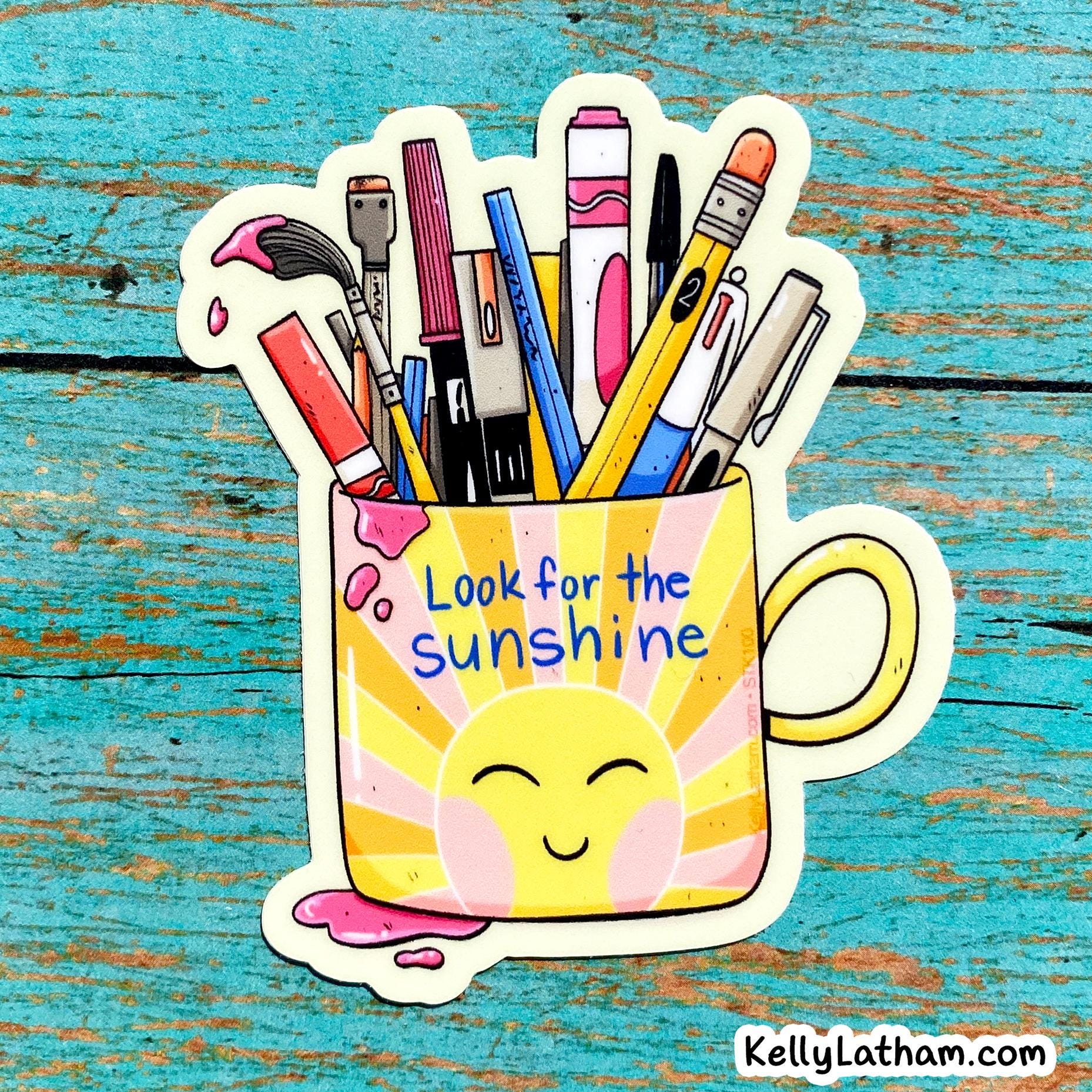 A die-cut sticker featuring a yellow and pink sun-faced mug filled with colorful art supplies, placed on a distressed turquoise wooden surface. The sticker reads 'Look for the sunshine' and includes a small 'Kellylatham.