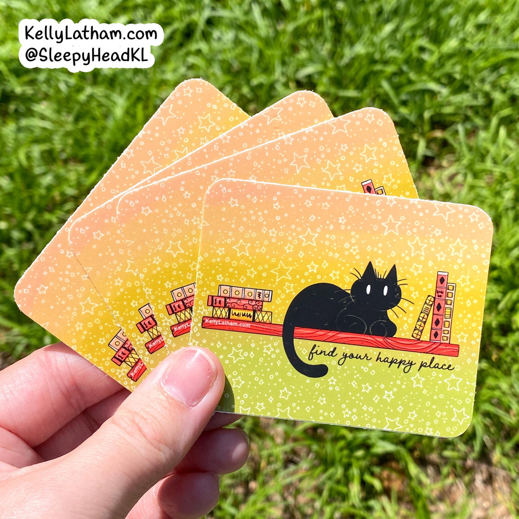 A hand holds four rectangular cards outdoors on grass. The cards have a yellow-to-green gradient background with white star speckles, a black cat illustration, and small red-and-white ticket graphics.