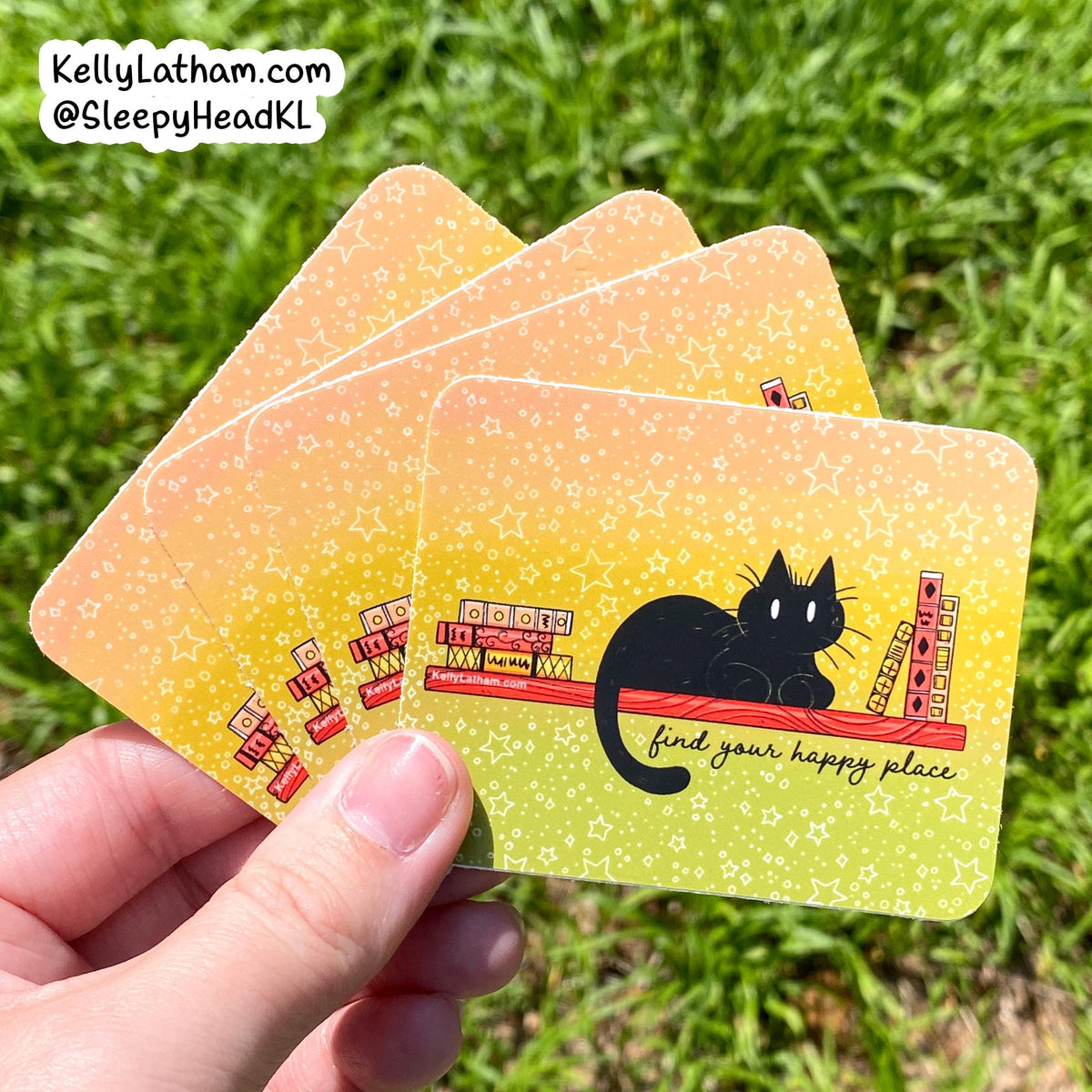 A hand holds four rectangular cards outdoors on grass. The cards have a yellow-to-green gradient background with white star speckles, a black cat illustration, and small red-and-white ticket graphics.