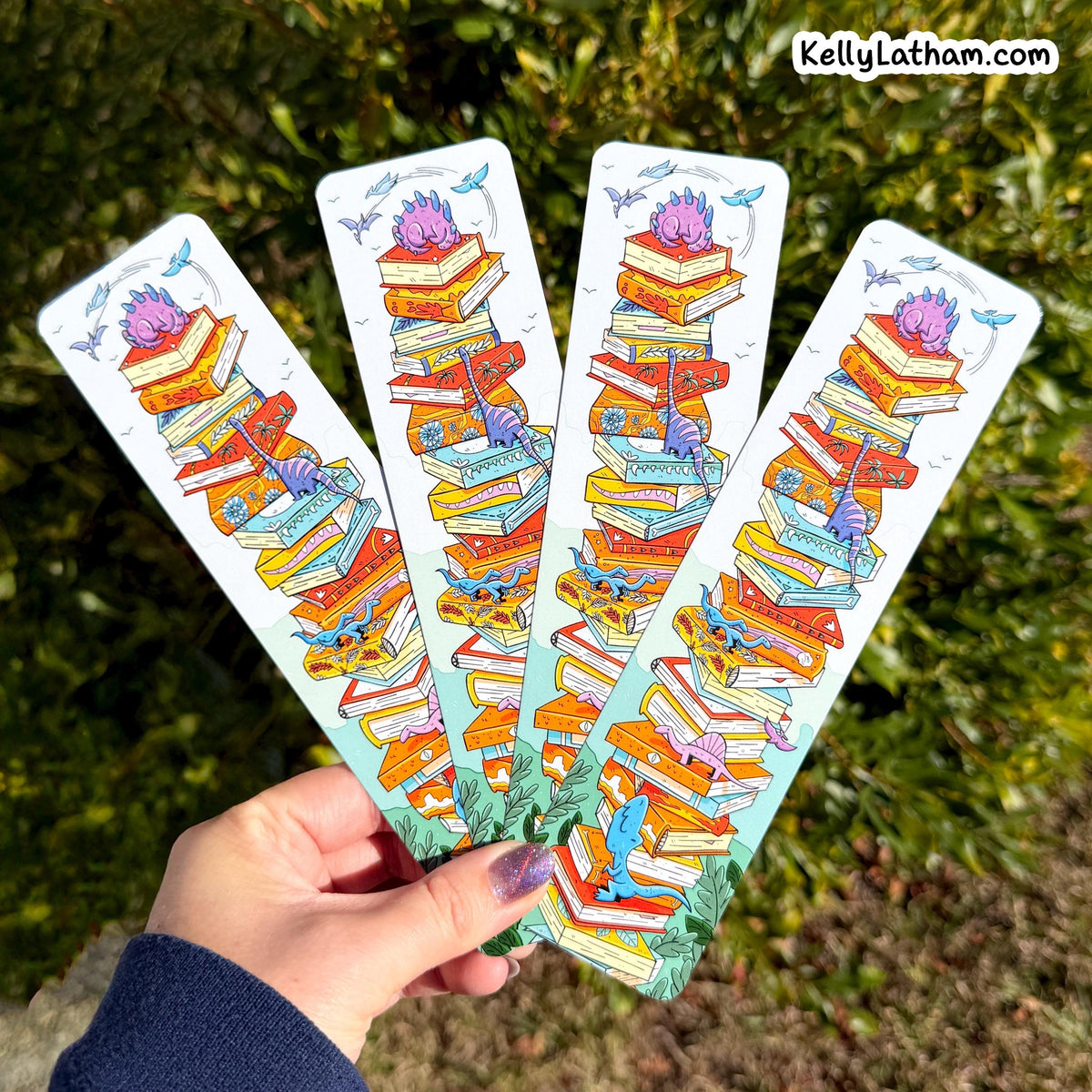 Four illustrated bookmarks are fanned out in a hand outdoors, with green foliage in the background. Each bookmark features a whimsical stack of colorful books, birds, and flowers.