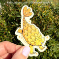 Hand holding a cartoon ankylosaurus sticker with green foliage in the background