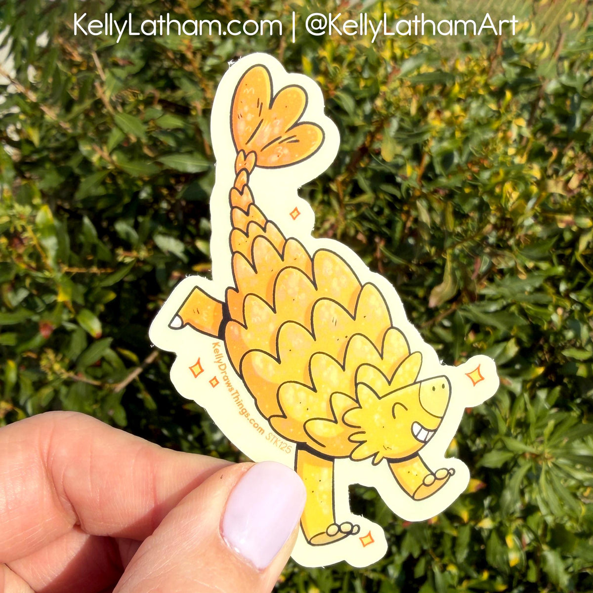 Hand holding a cartoon ankylosaurus sticker with green foliage in the background