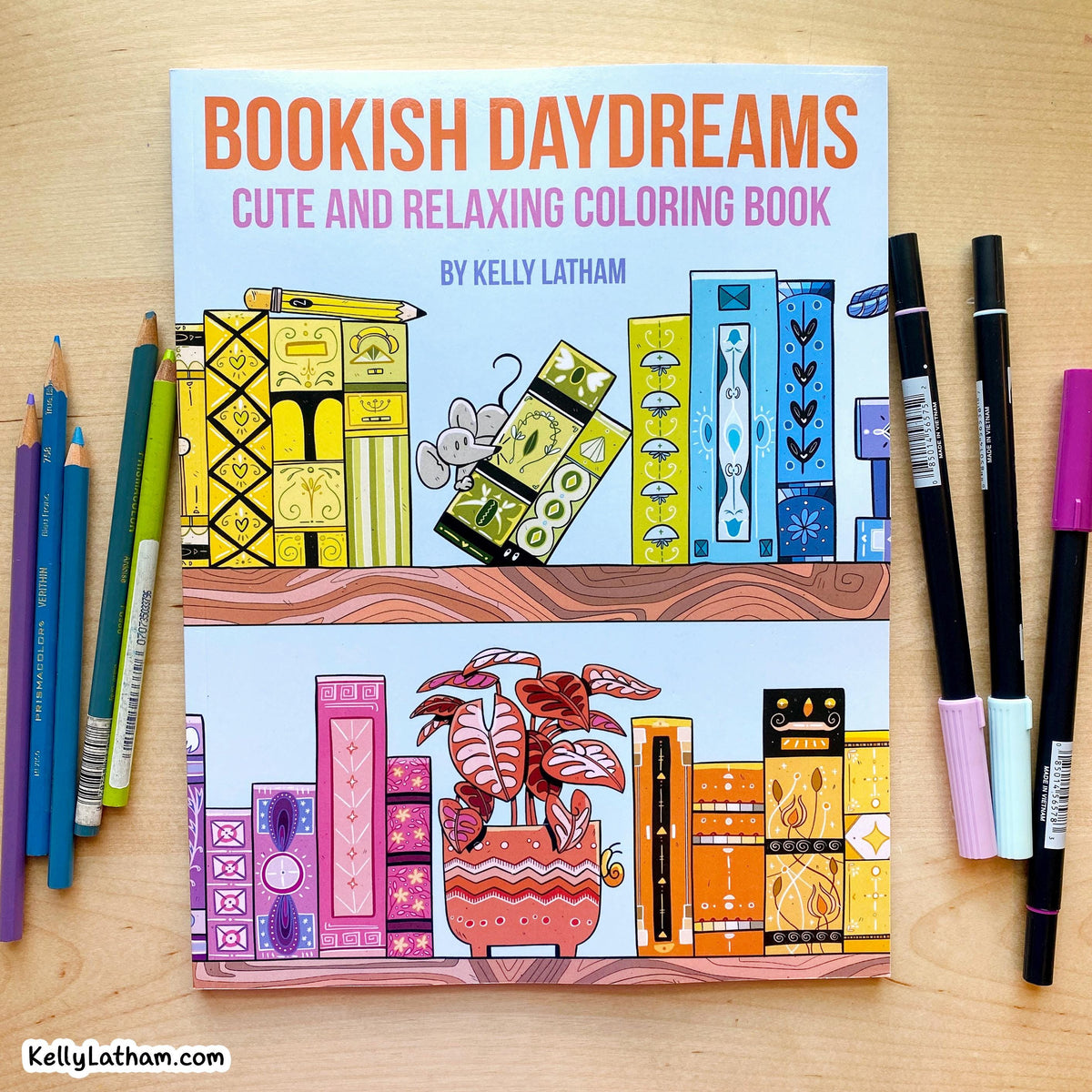 Bookish Daydreams: Cute and Relaxing Coloring Book by Kelly Latham, displayed on a light wood background with colored pencils on the left and markers on the right.