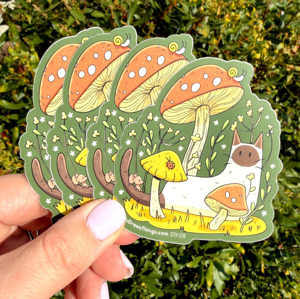 A hand holds a stack of die-cut stickers featuring a whimsical forest scene with large orange-and-white mushrooms, a white cat, and yellow flowers, photographed outdoors against green foliage.