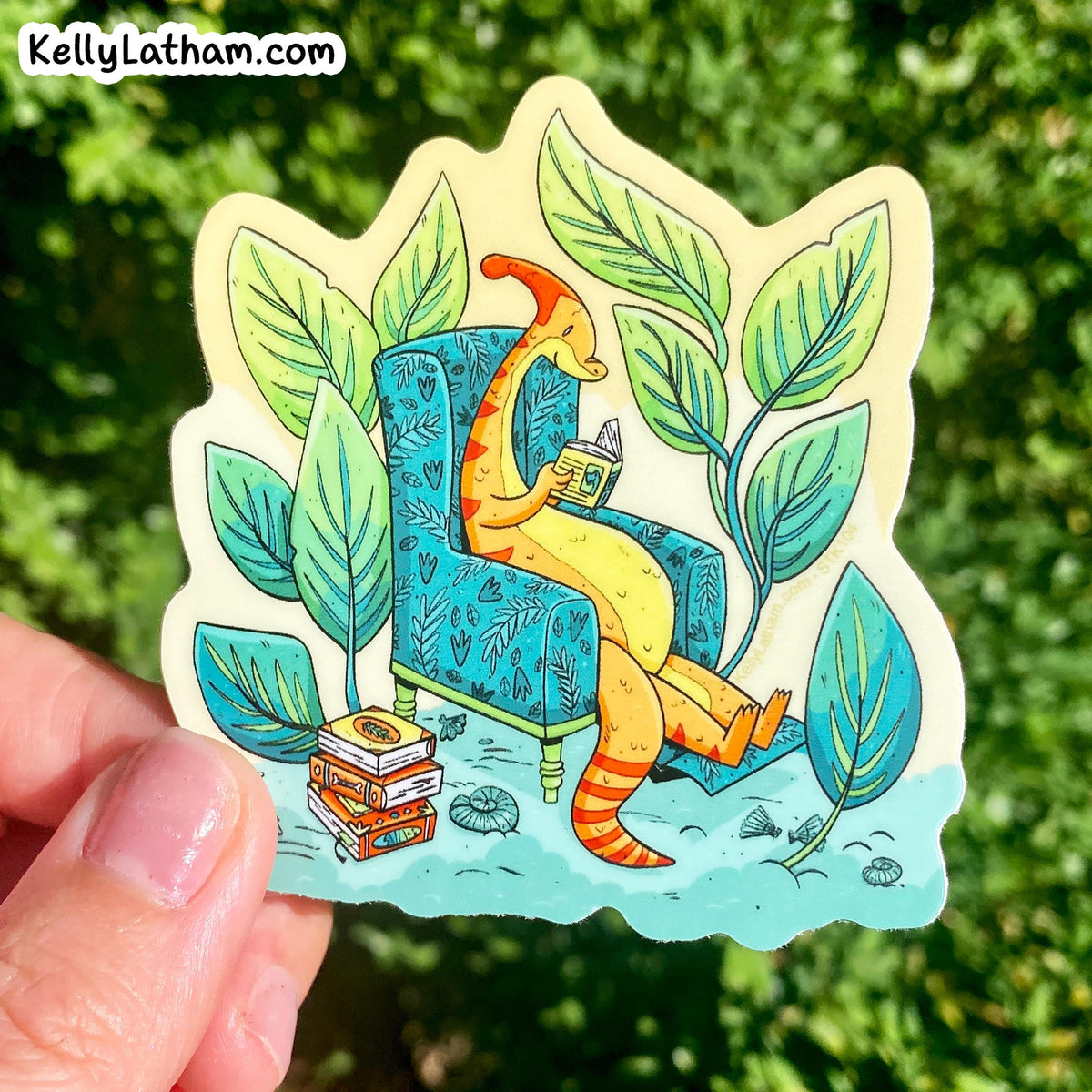 A hand holds a die-cut sticker featuring a whimsical illustration of a yellow-orange giraffe sitting in a blue patterned armchair and reading a book, surrounded by large green leaves, with a blurred green outdoor background.