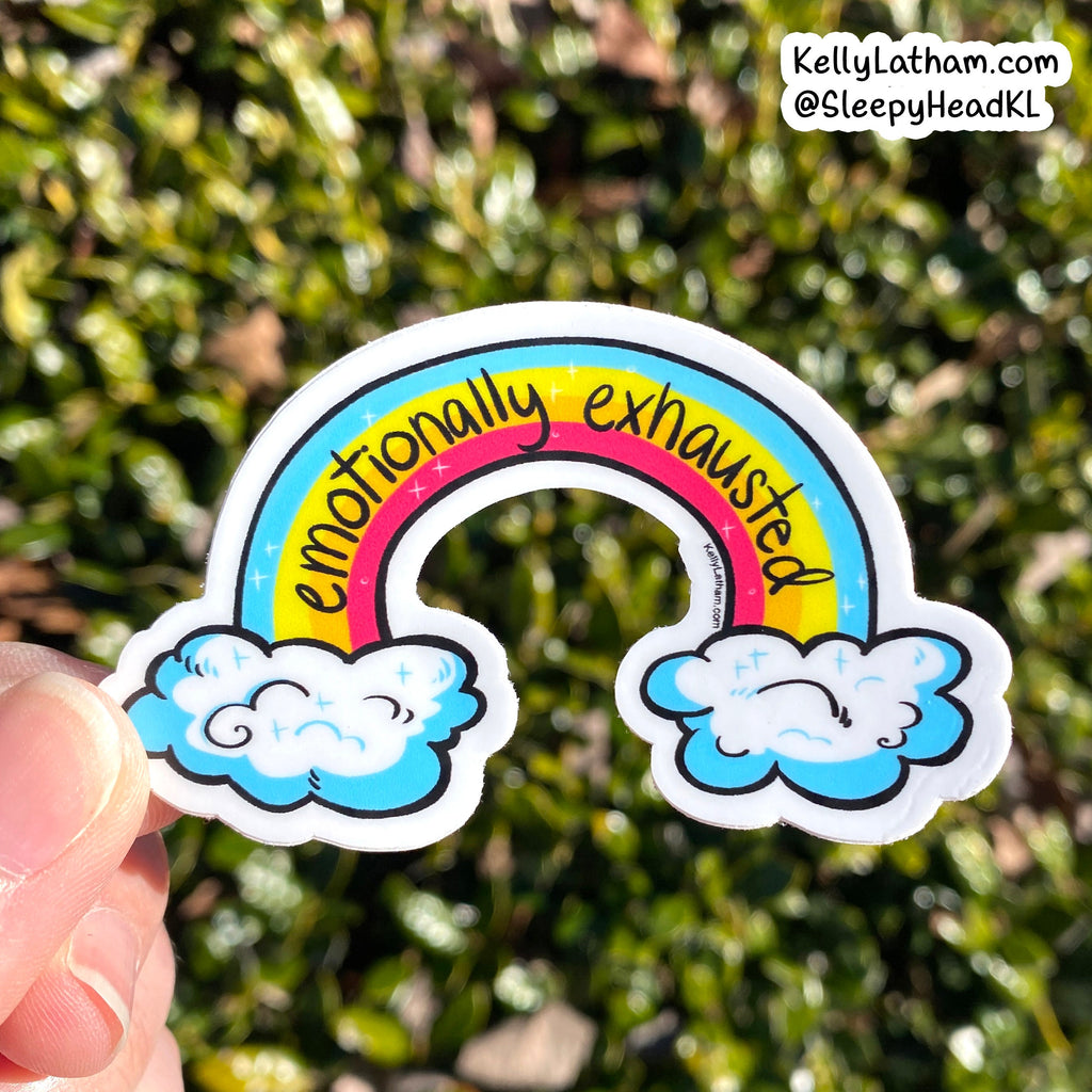 A hand holds a die-cut sticker featuring a cartoon rainbow with the words 'emotionally exhausted' in black script, set against a blurred green outdoor background. A brand watermark in the top right reads KellyLatham.