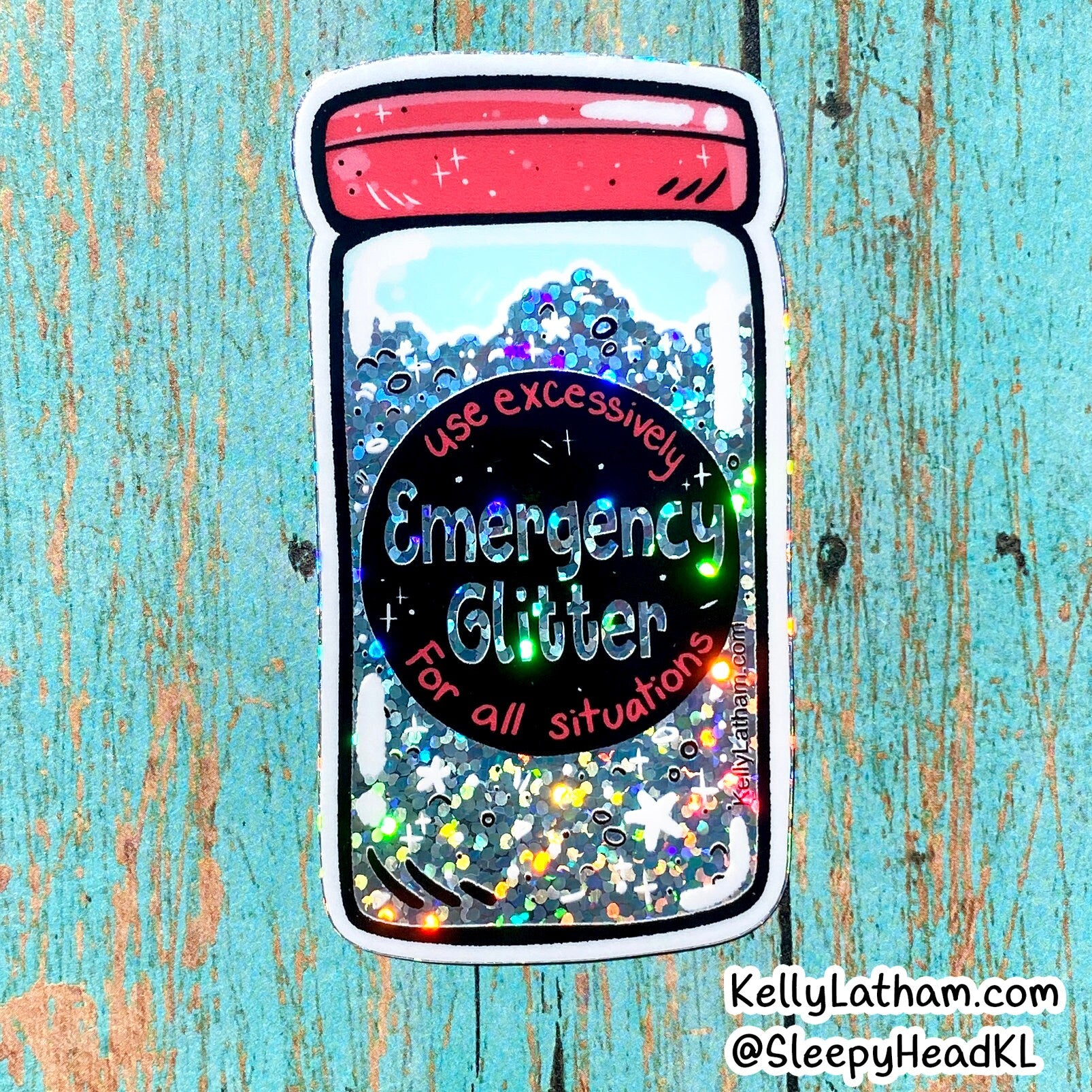 A die-cut sticker depicting a clear jar filled with holographic emergency glitter, featuring a black circular label that reads 'Emergency Glitter' and a red lid, shown on a distressed turquoise wood background.