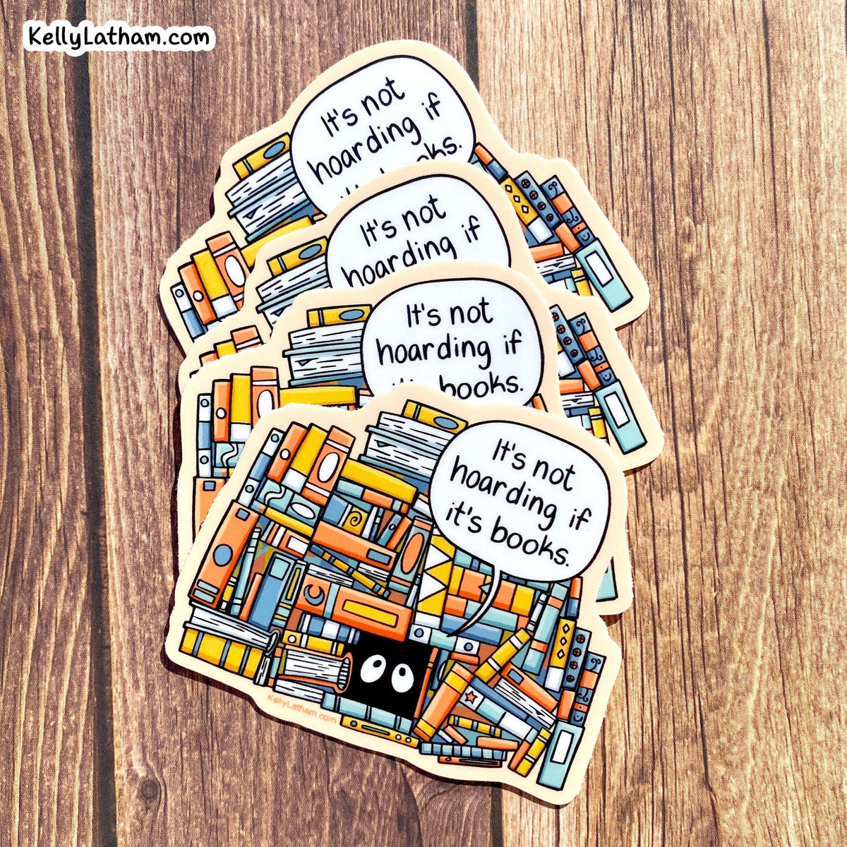 A set of die-cut stickers featuring a colorful illustration of books and three speech bubbles that read 'It's not hoarding if it's books.' The stickers are arranged in a fan shape on a wooden surface.