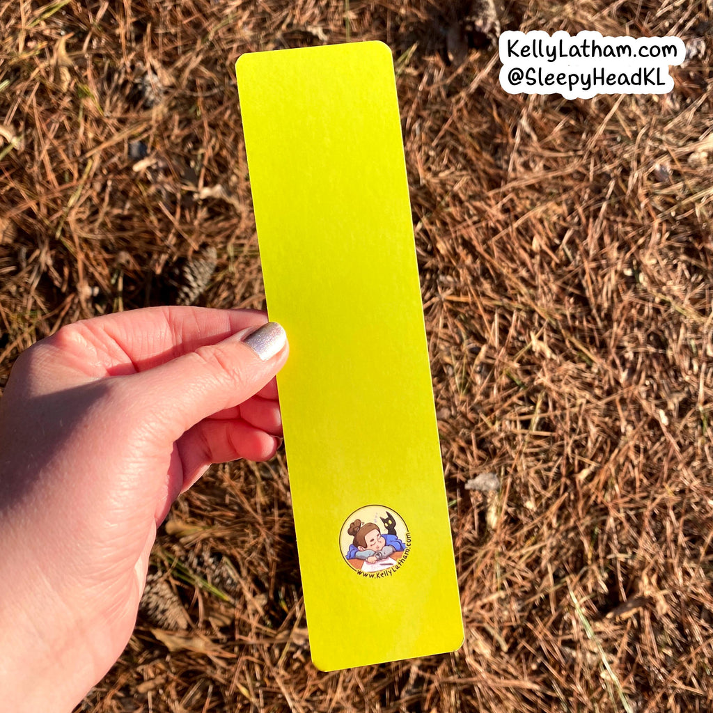 A hand holds a bright neon yellow bookmark outdoors against a background of dry pine needles. The bookmark features a small circular cartoon illustration near the bottom and has a white watermark in the top right corner.