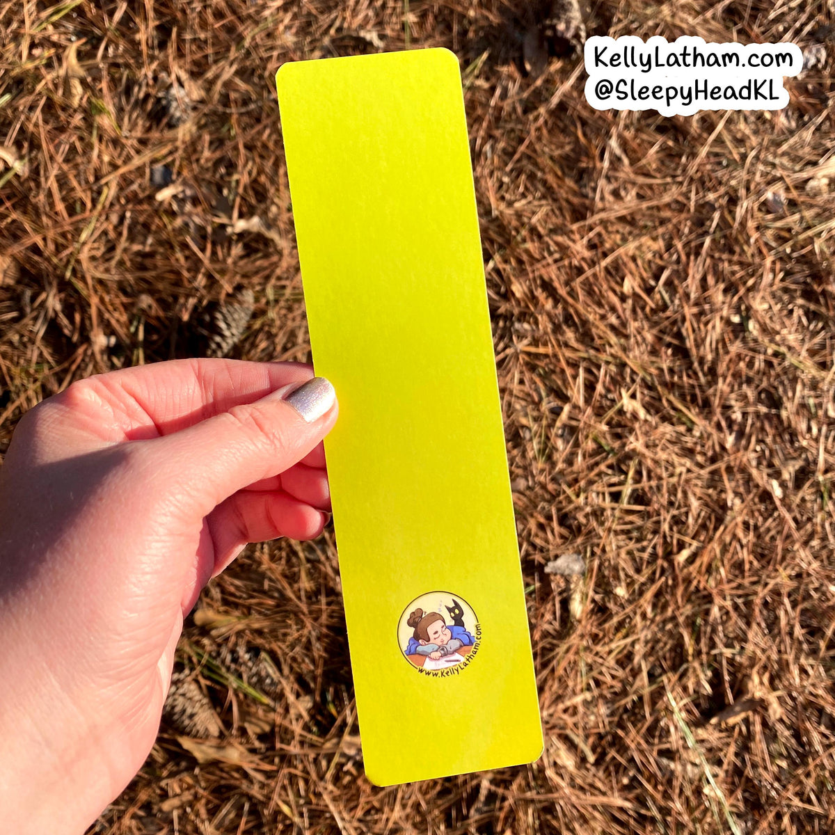 A hand holds a bright neon yellow bookmark outdoors against a background of dry pine needles. The bookmark features a small circular cartoon illustration near the bottom and has a white watermark in the top right corner.
