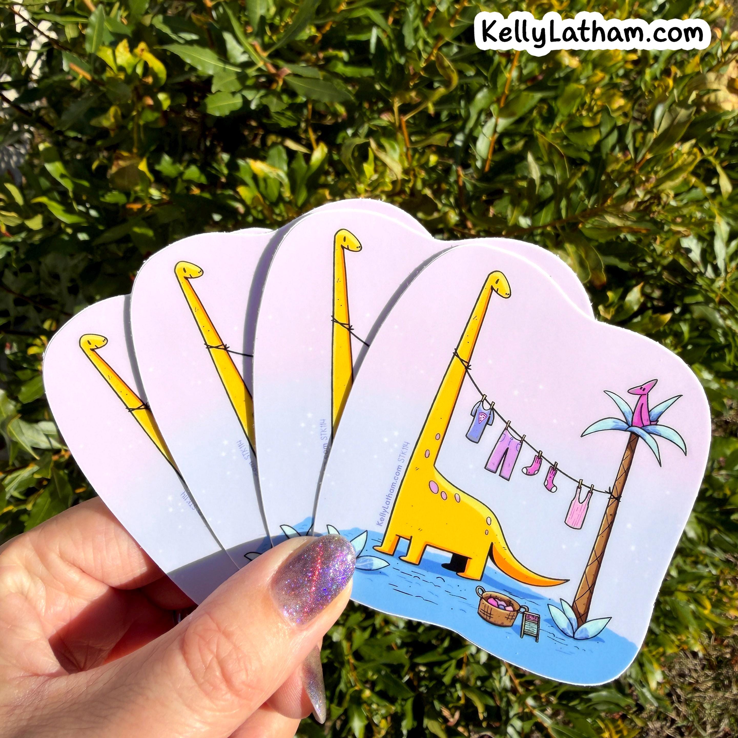 A hand holds a fan of four die-cut stickers outdoors against a background of green foliage. The stickers feature a whimsical illustration of a yellow long-neck dinosaur standing under a palm tree next to a clothesline with colorful laundry.