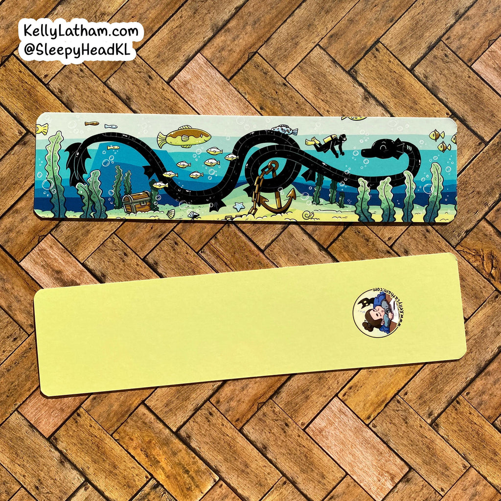 Two rectangular sleep masks laid flat on a wood herringbone floor. The top mask has an illustrated ocean scene with a large black serpent, blue water, fish, and sea plants.