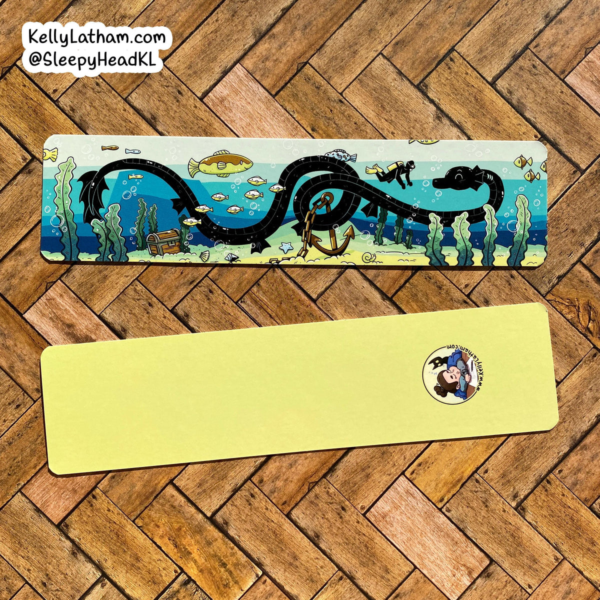 Two rectangular sleep masks laid flat on a wood herringbone floor. The top mask has an illustrated ocean scene with a large black serpent, blue water, fish, and sea plants.