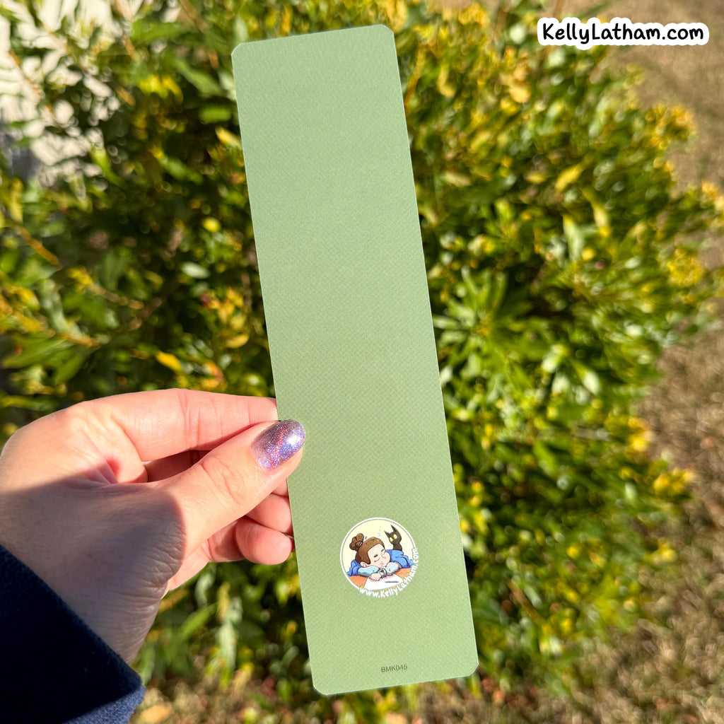 A hand holds a long, narrow sage green textured card outdoors in front of green foliage. The card has a small circular cartoon illustration near the bottom and the text 'KellyLatham.